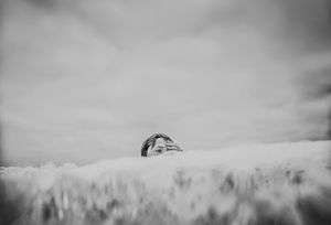 Black and white photo of a person's head barely visible above a field of tall grass under cloudy sky.