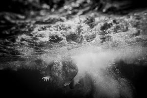 Dramatic black and white underwater photo of a person submerged beneath crashing ocean waves.