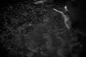 Black and white aerial view of a person swimming underwater with one hand reaching up through dark rippling water.