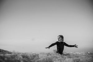 Joyful child splashing in ocean waves with arms outstretched, captured in black and white photography.