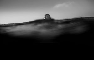 Black and white split-level photo of a person floating at the ocean surface, head barely visible above water.
