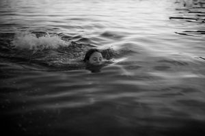 Black and white photo of a child swimming with head above calm dark water.