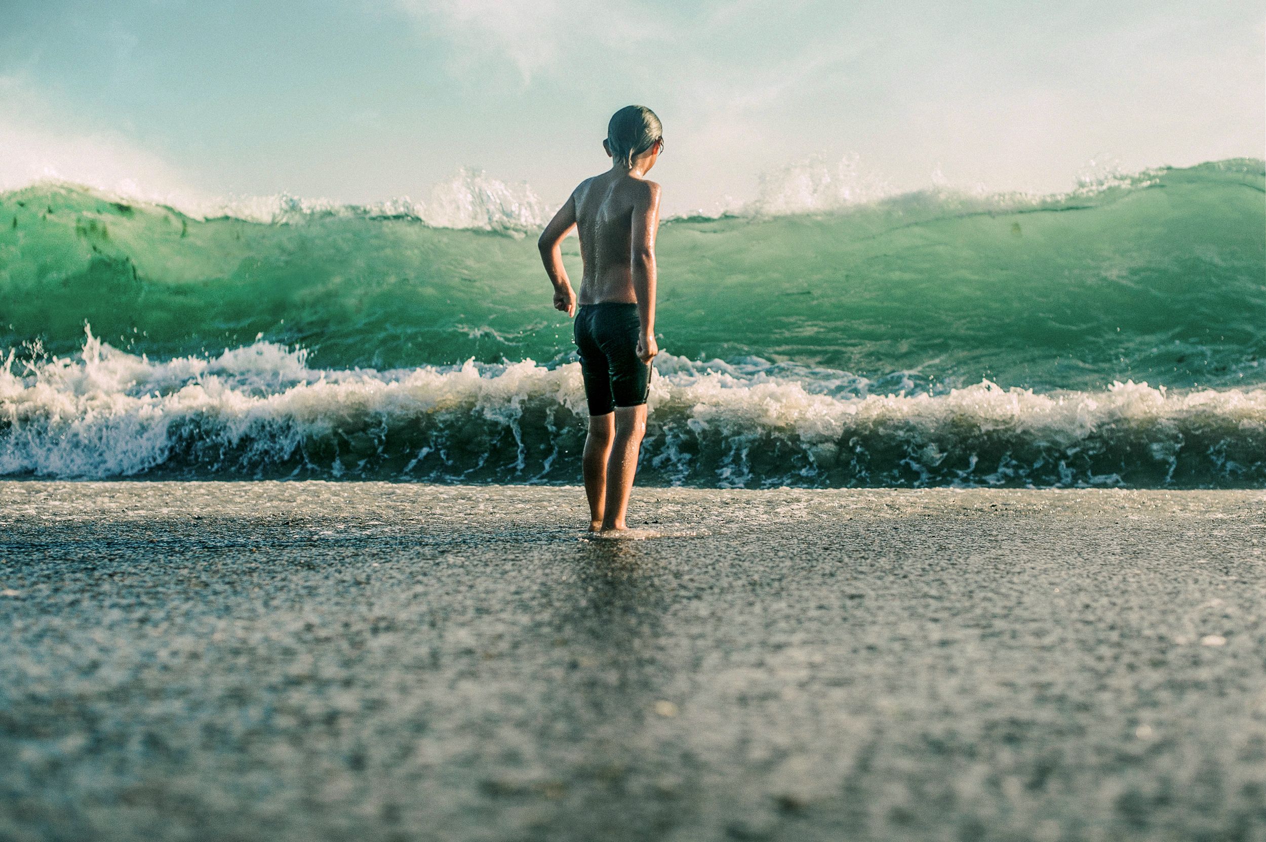 Young boy standing on a beach facing a large powerful green wave crashing toward the shore.