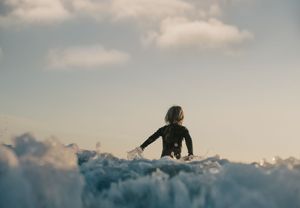 Young child in wetsuit sitting atop a foamy ocean wave under a cloudy sky.