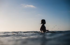 Silhouette of child sitting in calm ocean water at sunset.