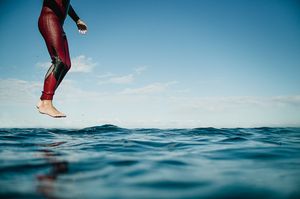 Surfer in red wetsuit standing on surfboard above calm blue ocean water.