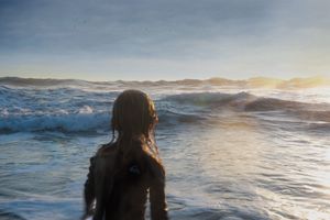 A child stands waist-deep in ocean waves at sunset, viewed from behind.