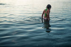 Young girl in green swimsuit wading in calm water at dusk.