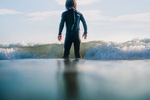 Surfer in full wetsuit standing on a surfboard facing an incoming ocean wave.