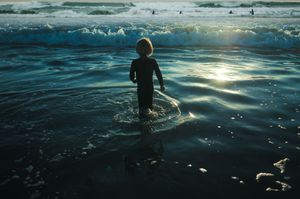 Silhouetted child in a wetsuit wading into the ocean at sunset with sparkling water reflections.