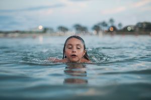 Young girl swimming in calm ocean water at dusk with bokeh lights in background.