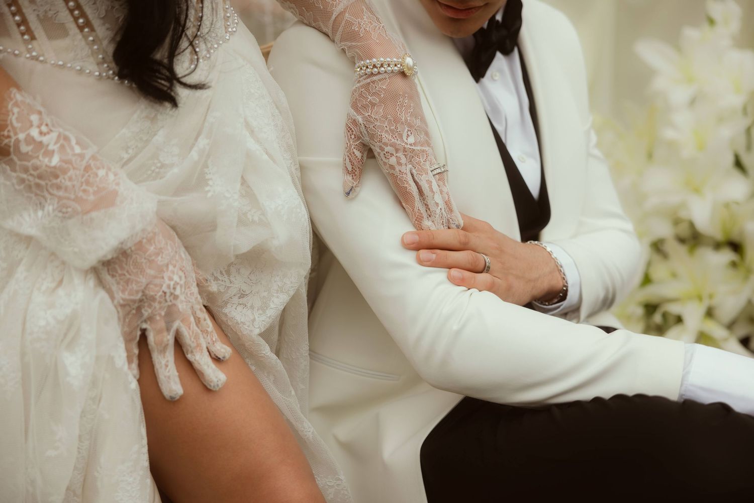 Bride in lace gloves and groom in white tuxedo sit together during elegant wedding ceremony.