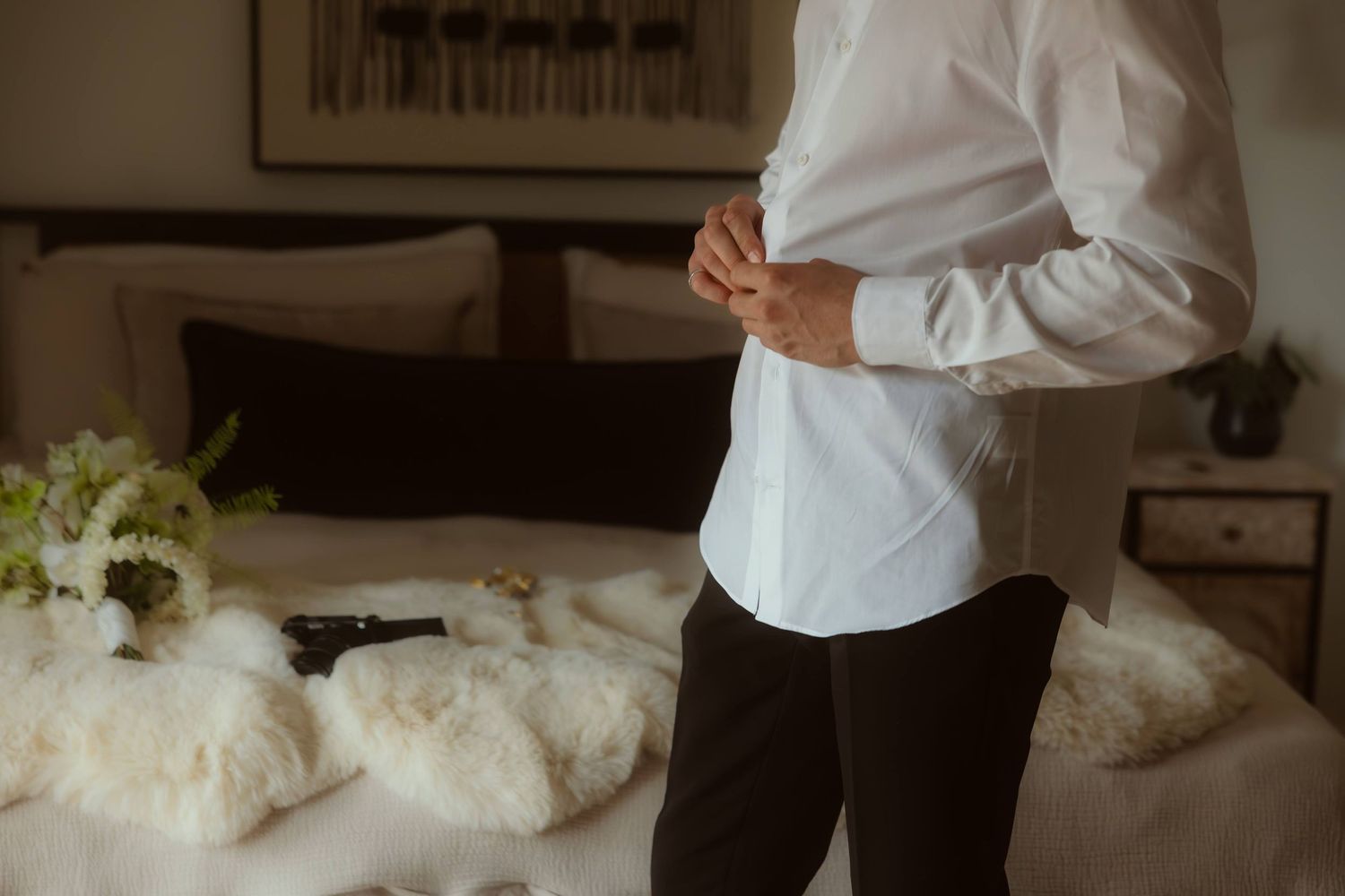 Groom in white dress shirt and black pants buttoning his shirt in a elegantly decorated bedroom.