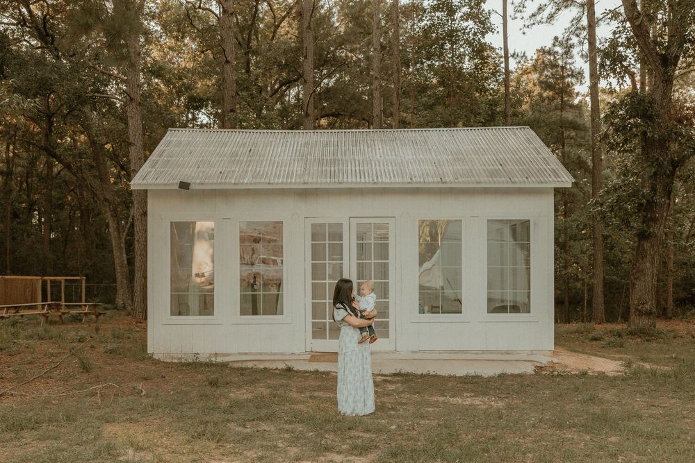 White wooden cottage in a forest setting with large windows and a rustic metal roof at sunset.