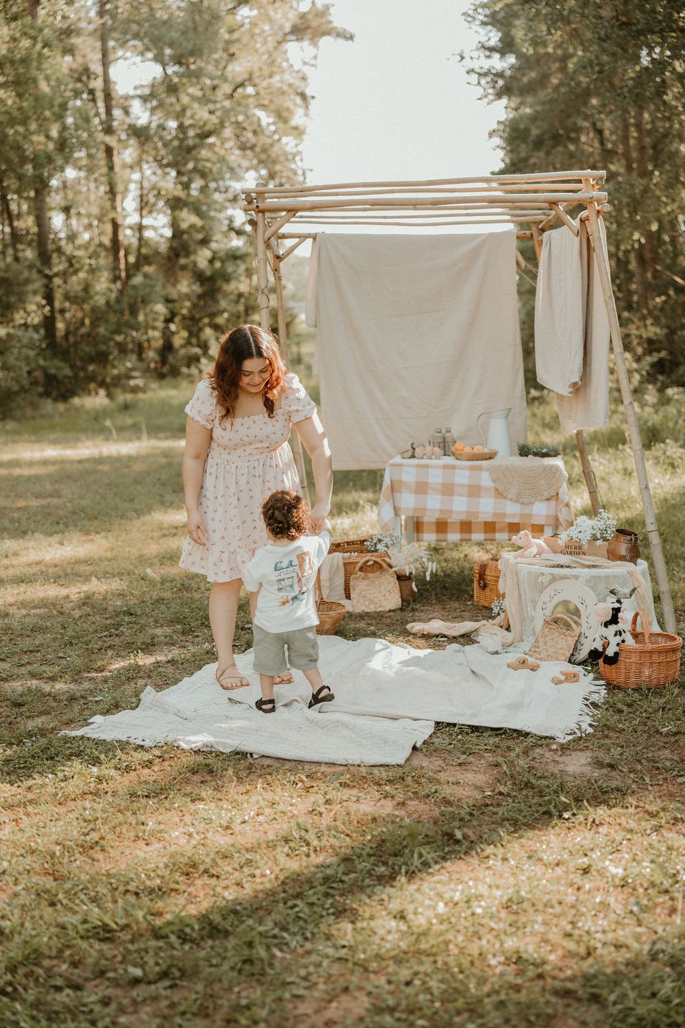 A rustic outdoor picnic setup with wooden frame backdrop, white blanket, and gingham tablecloth in a sunlit wooded setting.