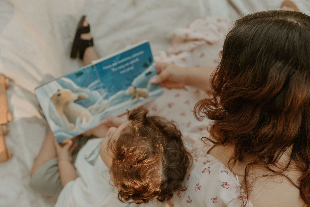 Two people lying on white bedding reading a children's book together during a cozy storytime moment.