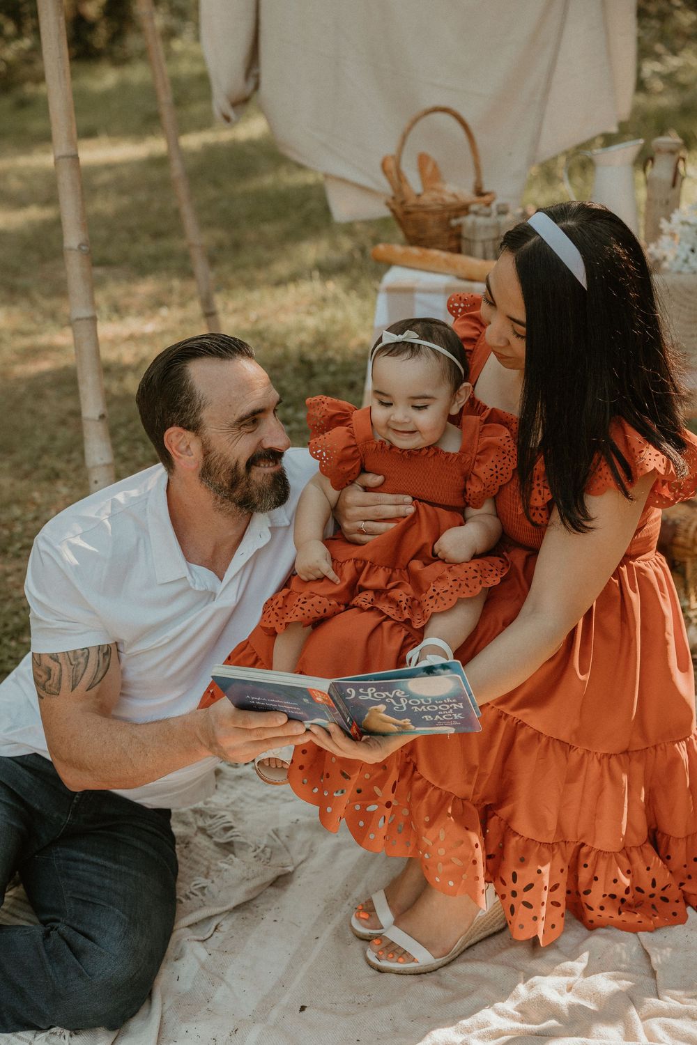 Family in matching orange outfits sits on blanket outdoors while looking at picture book together in warm sunlight.
