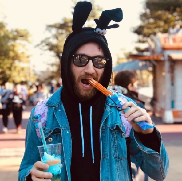 Bearded man wearing bunny ears hoodie and sunglasses eating a churro at an amusement park.