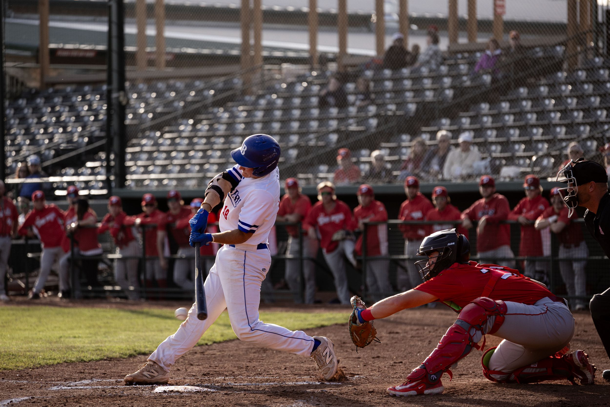 Brooks Bombers (WCBL) batter swings at pitch with catcher and umpire behind home plate at sunset.