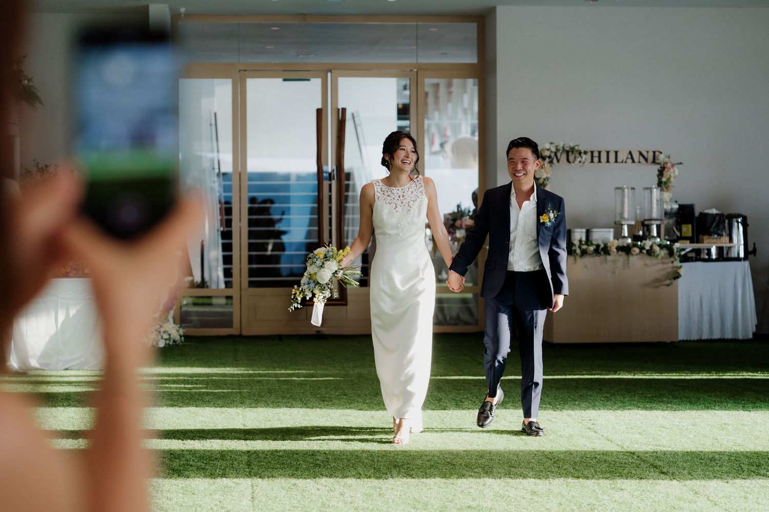 A bride and groom walk hand-in-hand at their wedding reception on a green lawn, as a guest captures the moment on a phone.