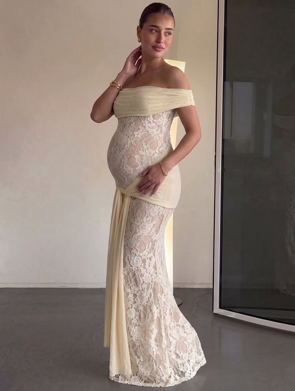Pregnant woman in elegant off-shoulder cream lace mermaid gown with ruched bodice and flowing train.