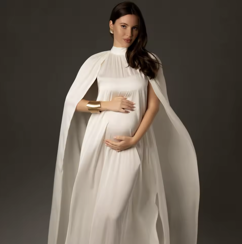 Pregnant woman in elegant white cape maternity gown with gold cuff bracelet poses gracefully against gray background.