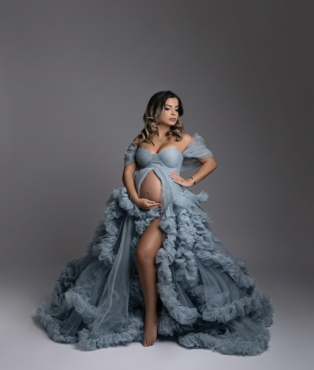 Pregnant woman in elegant dusty blue ruffled tulle maternity gown posing gracefully in professional studio photo.