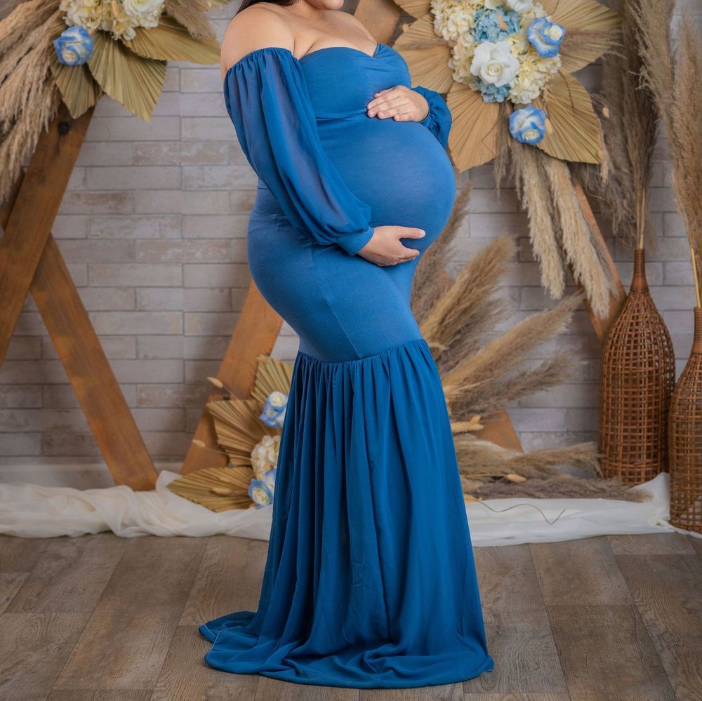 Pregnant woman in elegant blue off-shoulder maternity gown with long sleeves and mermaid skirt, posing in boho studio.