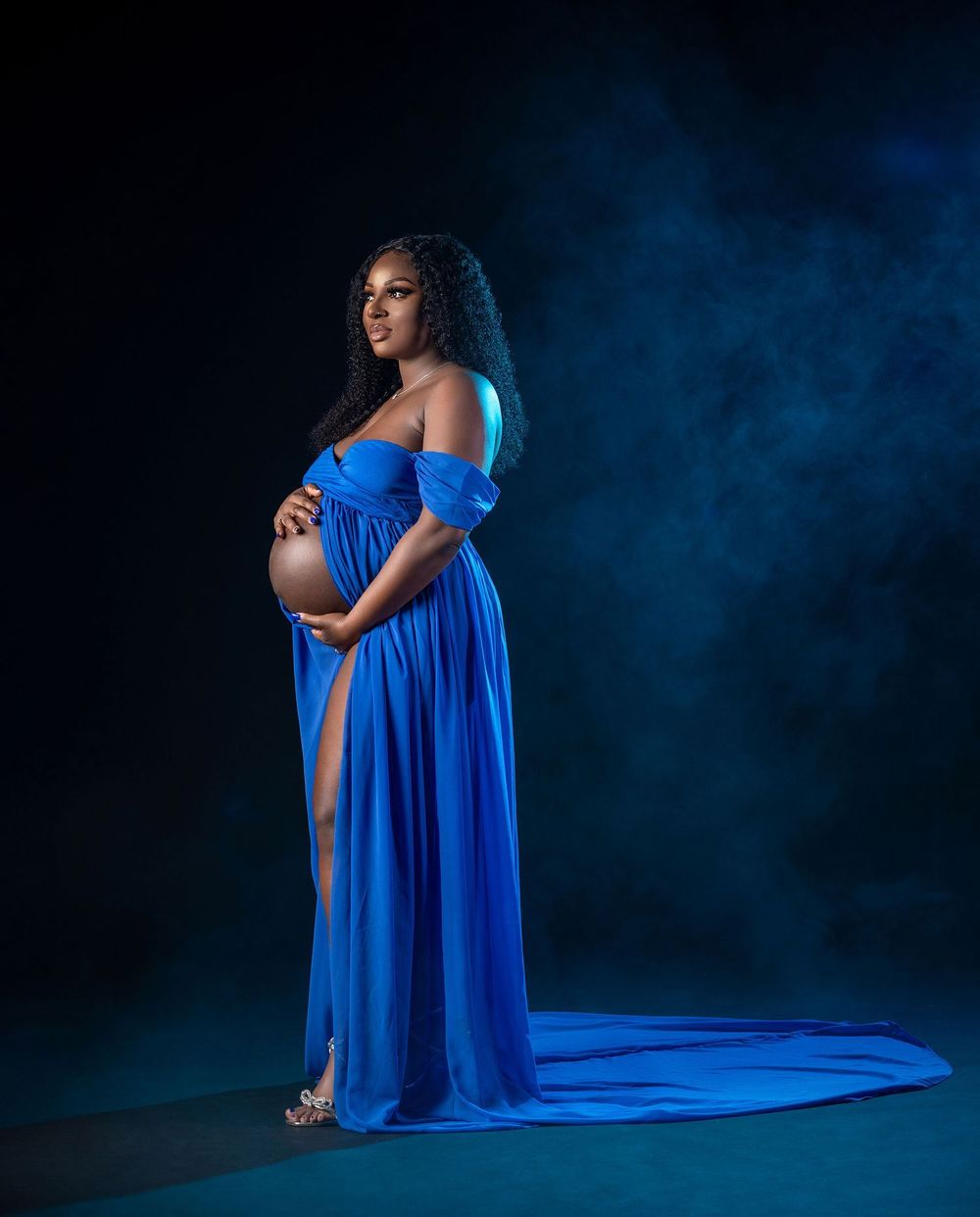 Pregnant woman in elegant royal blue off-shoulder maternity gown posing gracefully in a dramatic studio setting.