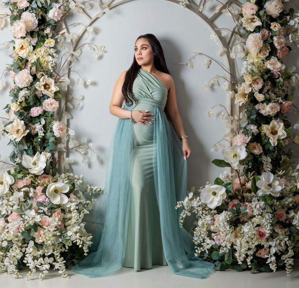 Pregnant woman in sage green one-shoulder mermaid gown with tulle overlay, posing before a floral arch backdrop.