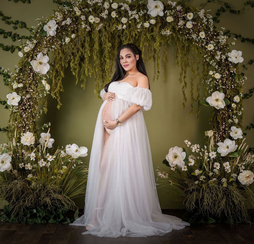 Pregnant woman in white off-shoulder gown poses under floral arch with white blooms and greenery.