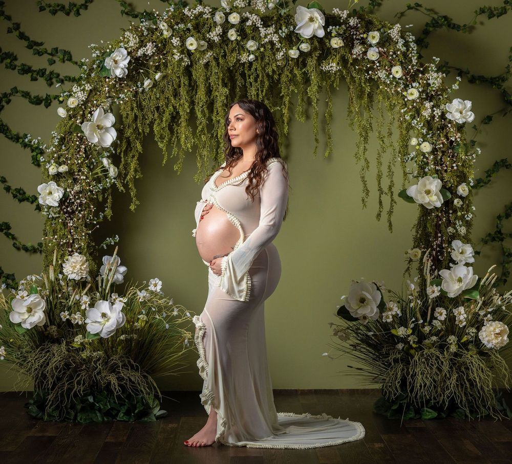 Pregnant woman in white gown poses under floral arch with white flowers and greenery on green backdrop.