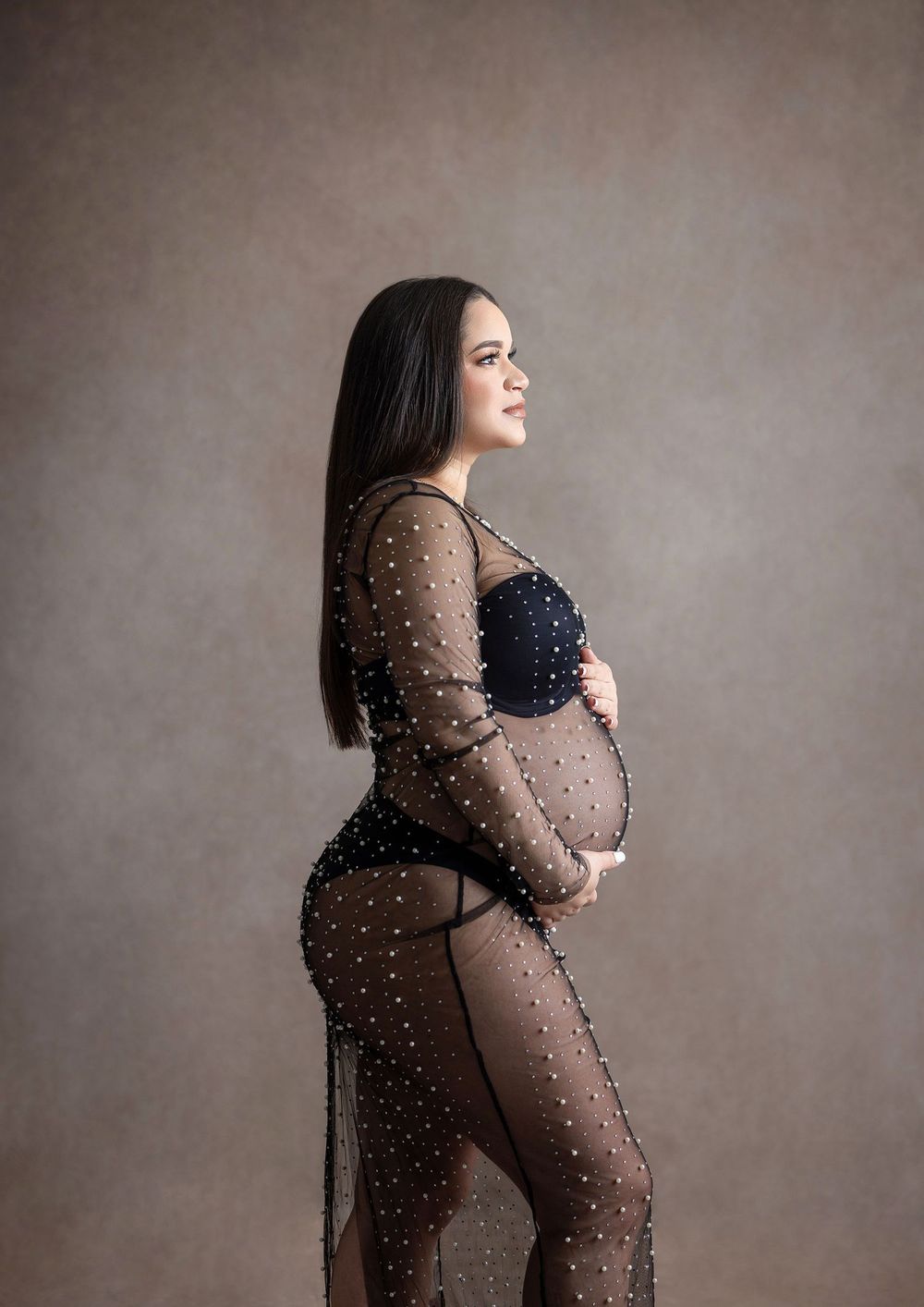 Pregnant woman in black sheer embellished maternity gown posing sideways in professional studio photo shoot.