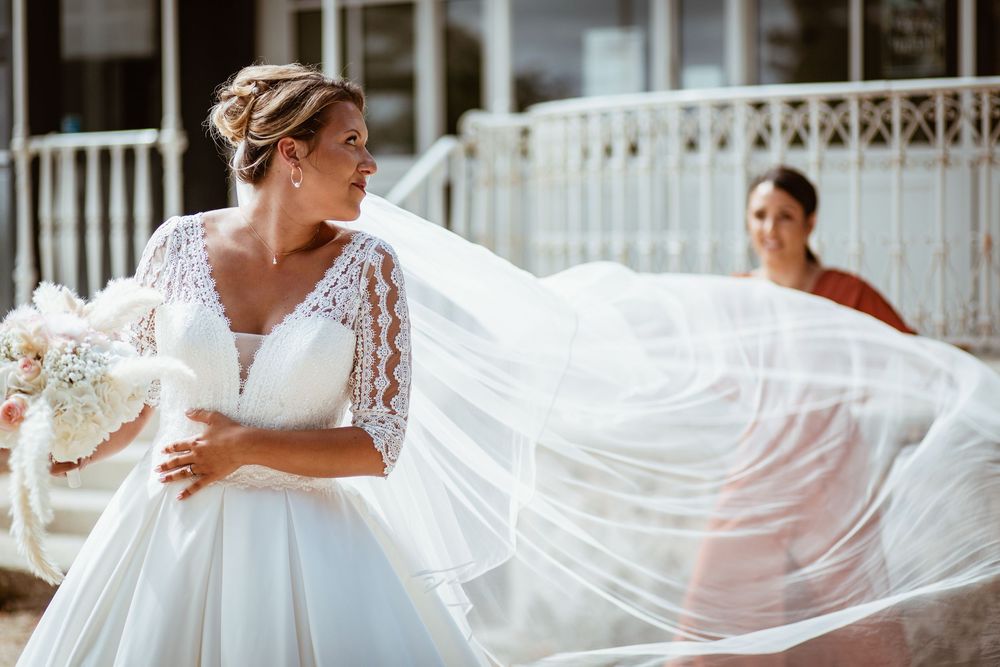 Bride in lace wedding dress with flowing veil holding bouquet, bridesmaid in red dress visible in background.