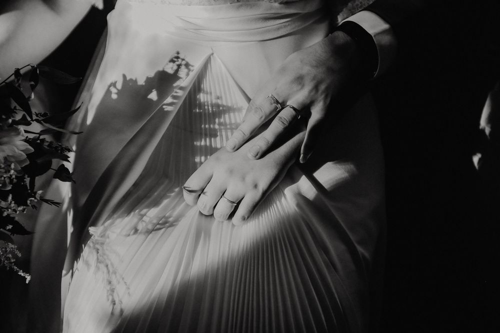 Close-up black and white photo of bride and groom hands with wedding rings on pleated wedding dress.