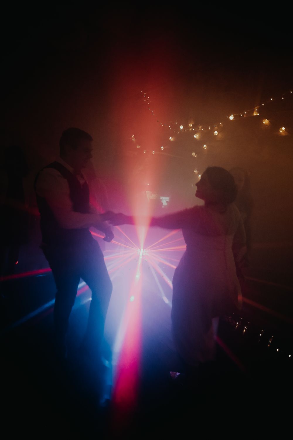 Bride and groom dancing under colorful laser lights and string lights at wedding reception.