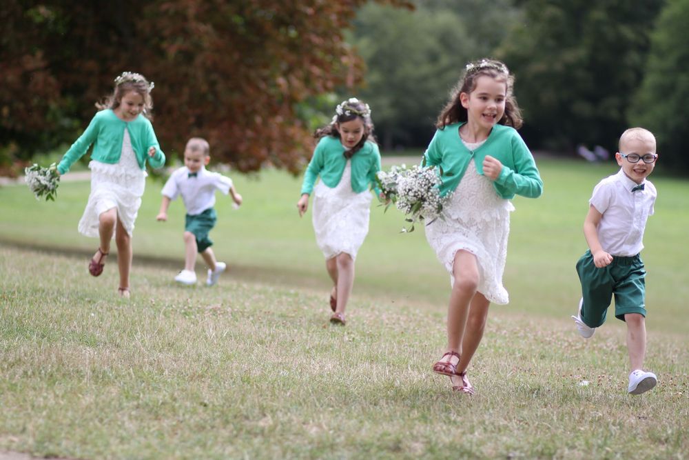 Children in white dresses and green cardigans run joyfully across a grassy field holding bouquets at a wedding.