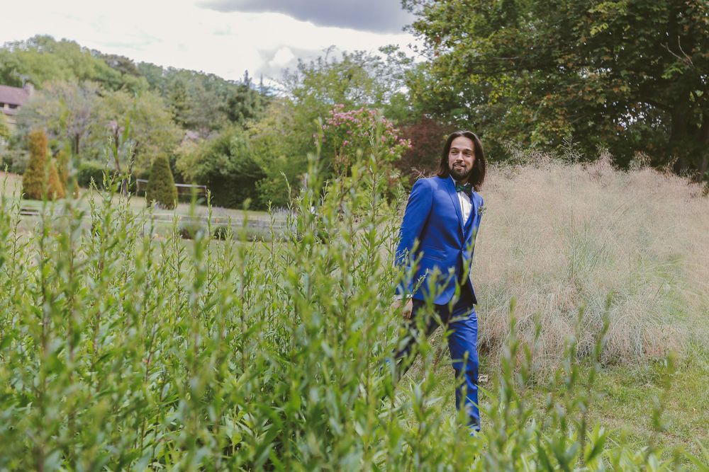 Woman in blue outfit standing among lush green plants in a garden with trees and hills in the background.