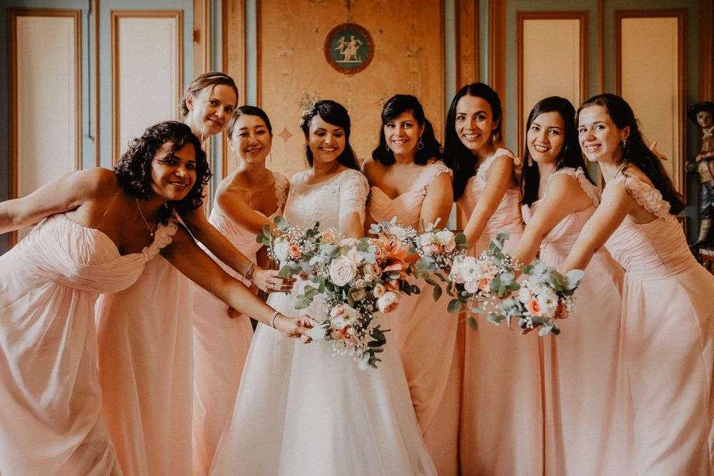 Bride in white gown poses with seven bridesmaids in blush pink dresses holding floral bouquets indoors.