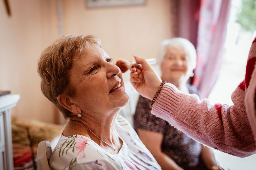 Elderly woman having makeup applied while another senior woman smiles in background.
