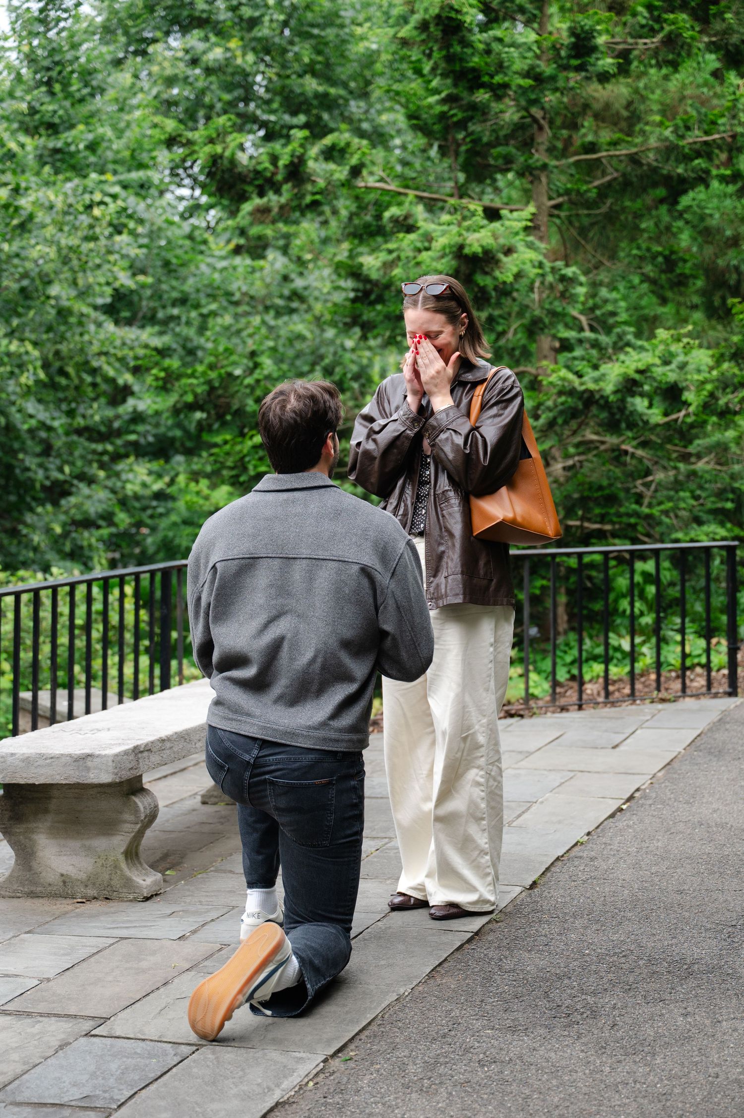 Man proposing on one knee to emotional woman in a park, with lush green trees in the background.