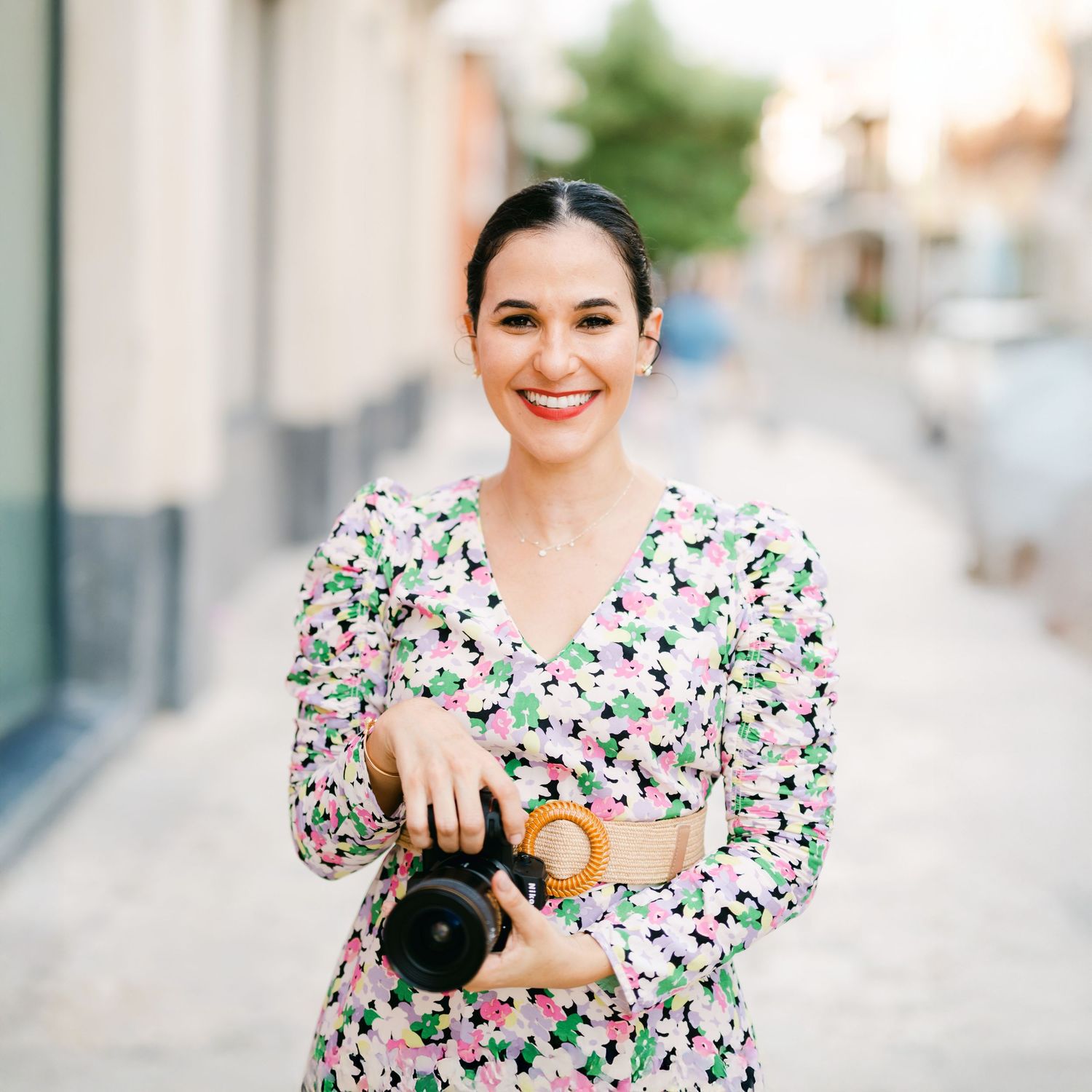Photographer in floral dress poses with camera outdoors in bright natural lighting.