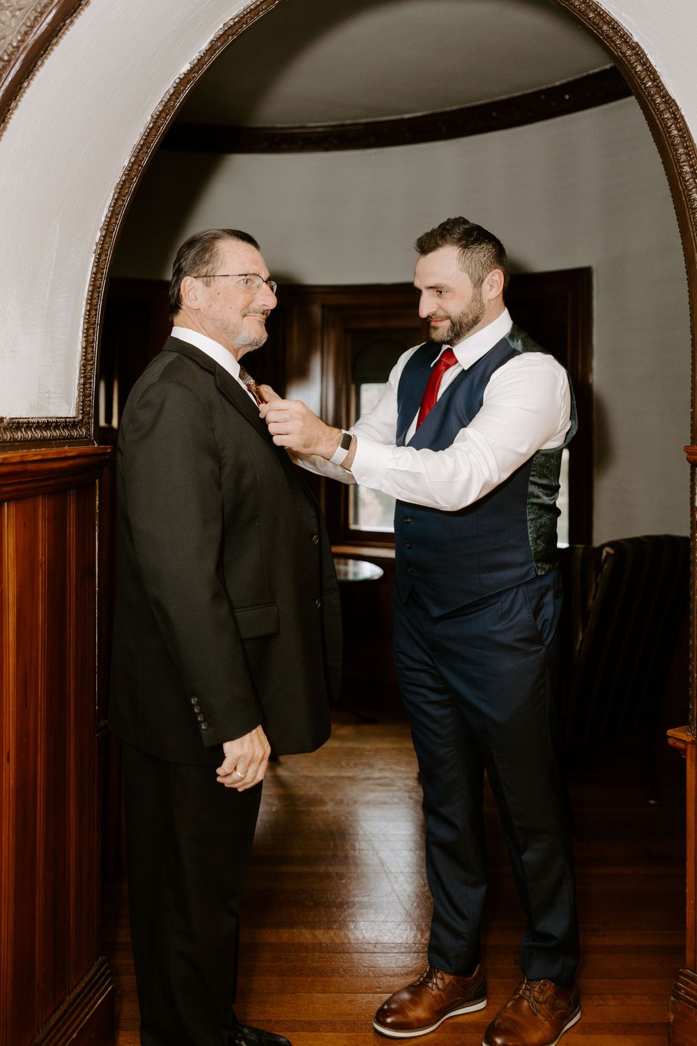 Groom in navy suit adjusting older mans tie under an elegant arched doorway indoors.