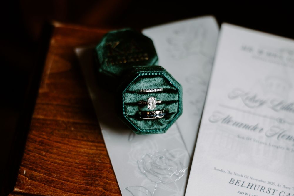 Diamond engagement ring and wedding bands displayed in a green velvet ring box with invitation.