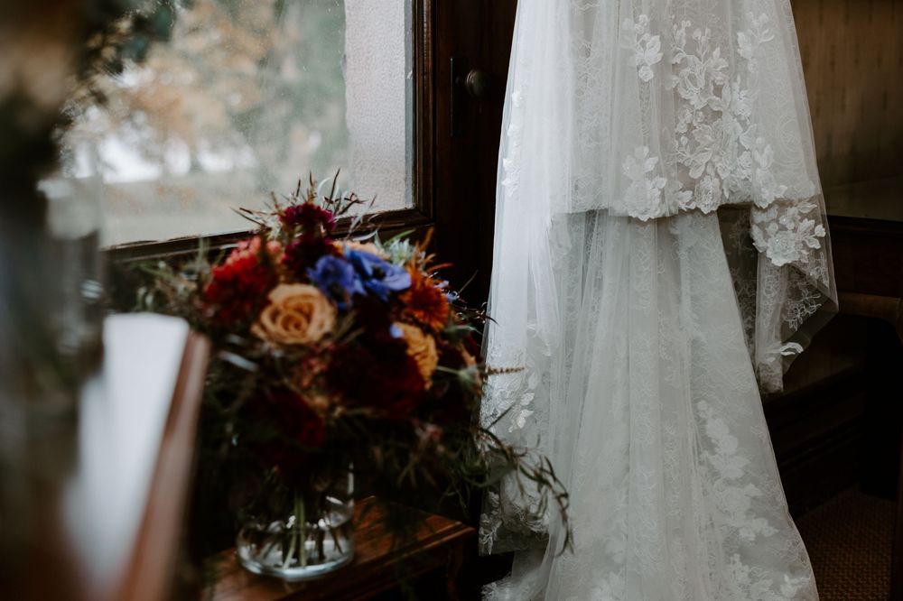 Lace wedding dress hanging by a window next to a vibrant bouquet of red, blue, and orange flowers.