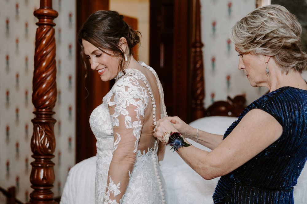 Mother helping bride button up her lace wedding gown before the ceremony.