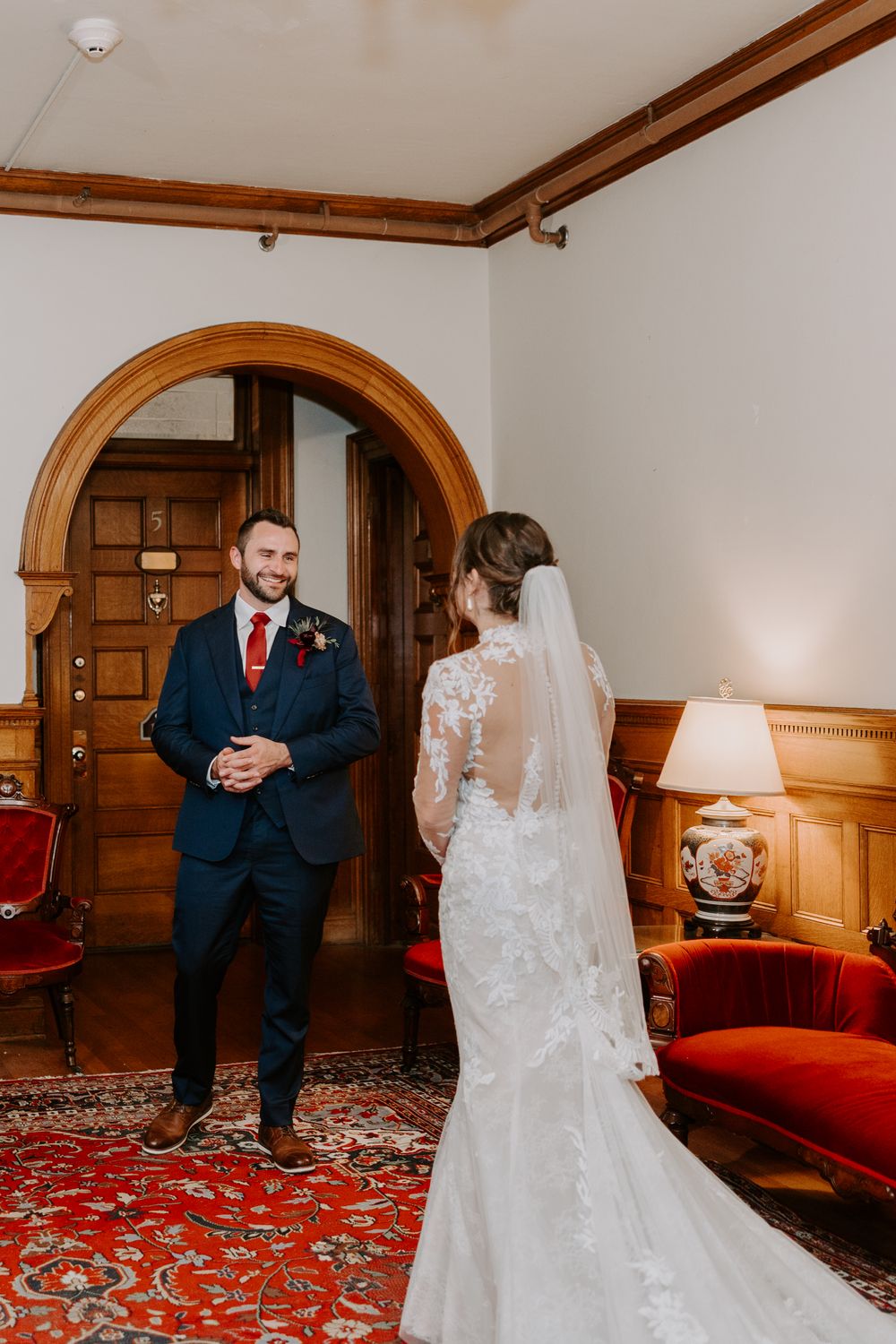 Groom in navy suit smiling at bride during first look in a classic interior.