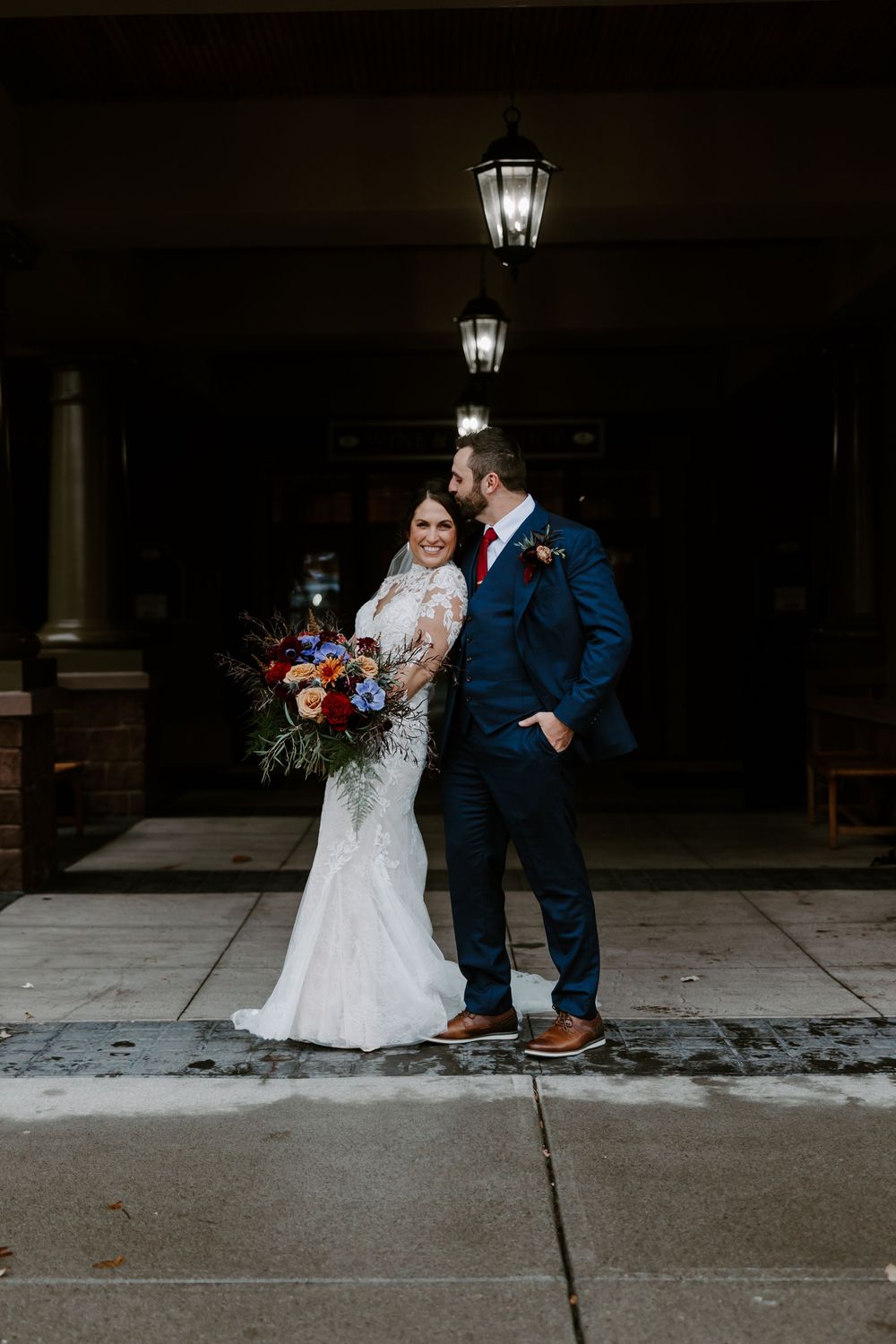 Bride and groom posing under lanterns outdoors with a vibrant fall floral bouquet.