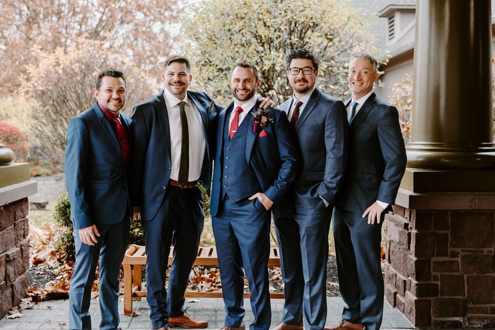 Groom and groomsmen in navy suits smiling together outdoors in autumn setting.
