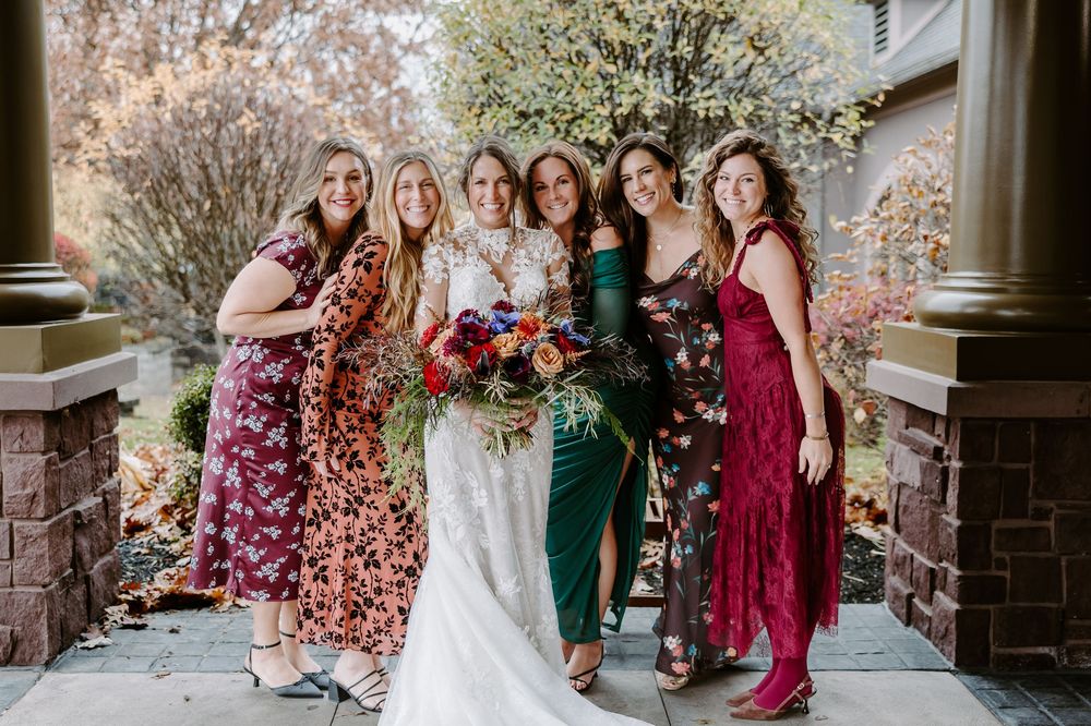 Bride poses with five bridesmaids in colorful floral dresses holding a vibrant bouquet outside a brick venue.